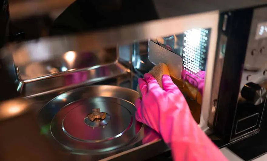 Een roze handschoen maakt een zilveren microgolfoven schoon met een gele spons.