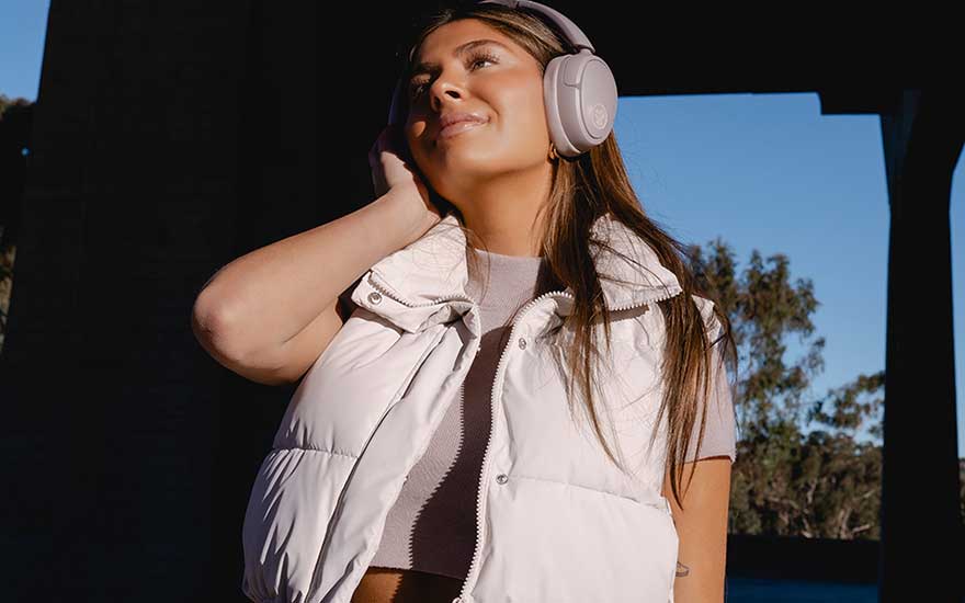 Une personne portant un casque audio blanc et un gilet léger écoute de la musique en extérieur par une journée ensoleillée, sous un ciel bleu.