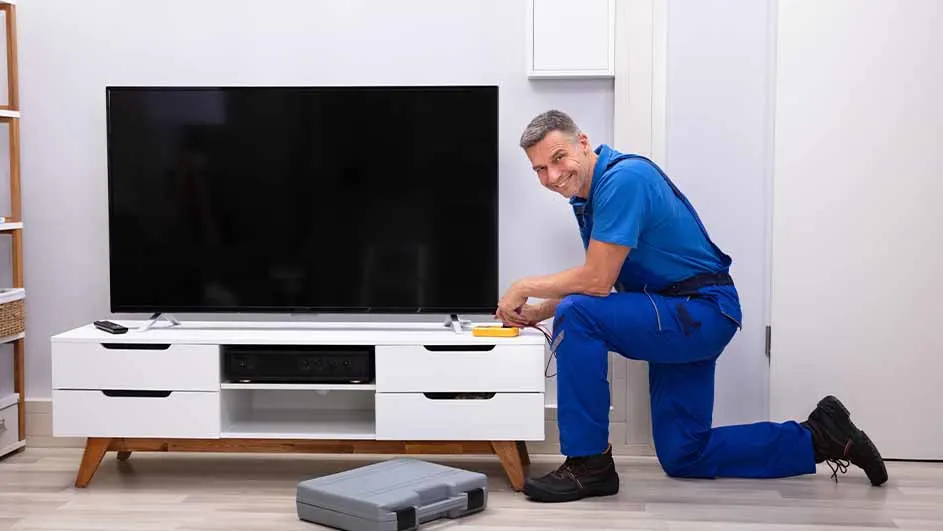 Un technicien en bleu travaille sur un téléviseur noir sur un meuble TV blanc. Une boîte à outils grise est au sol.