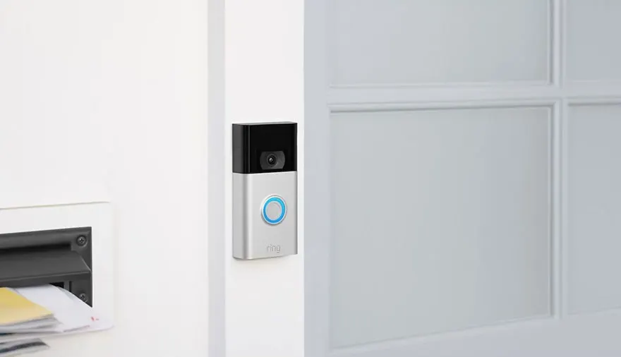 Une sonnette vidéo argentée avec un bouton bleu est fixée sur un mur blanc à côté d'une porte blanche.