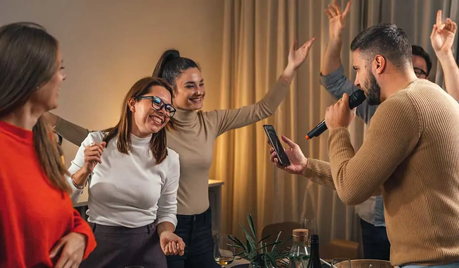 Des gens à une fête chantent du karaoké avec un micro et un téléphone devant des rideaux beige. Des bouteilles et des verres sont sur une table.