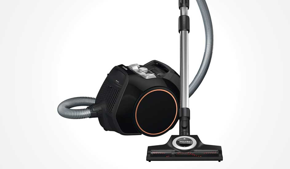 Black cylinder vacuum with orange trim, flexible hose and metal tube ending in wide floor head, shown against a light background.