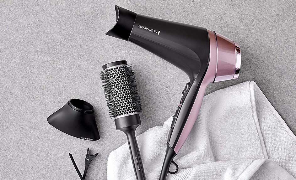Een zwarte en roze Remington haardroger, een ronde borstel, een mondstuk en haarclips op een grijze achtergrond met een witte handdoek.