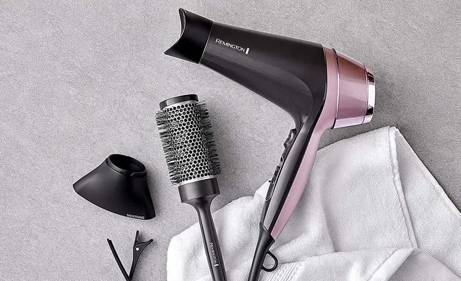 Een zwarte en roze Remington haardroger, een ronde borstel, een mondstuk en haarclips op een grijze achtergrond met een witte handdoek.