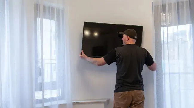 A black TV is mounted on a white wall, between two windows covered with white curtains.
