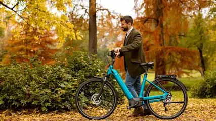 Un homme souriant tient un vélo bleu sur un chemin couvert de feuilles d'automne, entouré de buissons et d'arbres aux feuilles jaunes et orange.