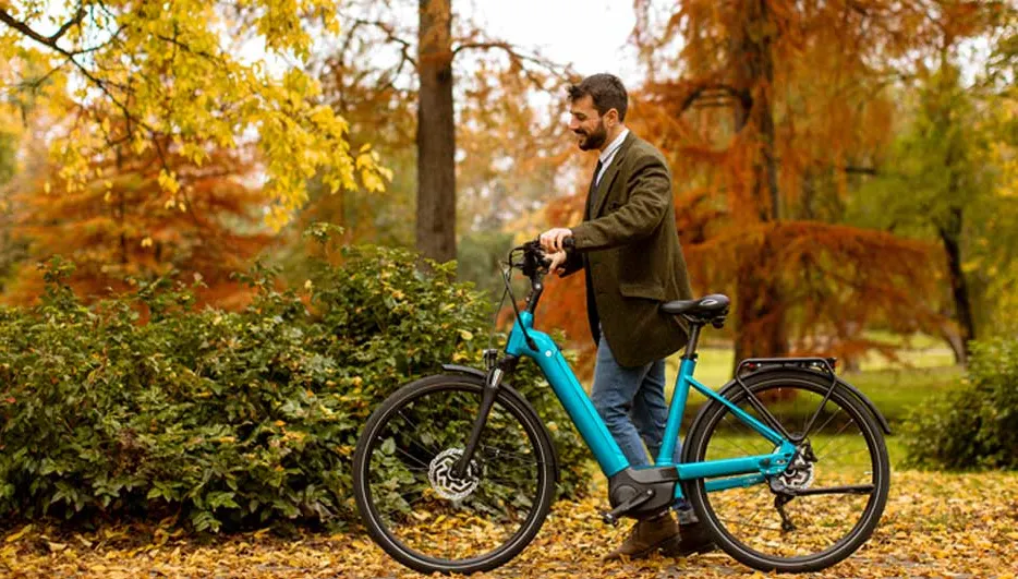 Un homme souriant tient un vélo bleu sur un chemin couvert de feuilles d'automne, entouré de buissons et d'arbres aux feuilles jaunes et orange.