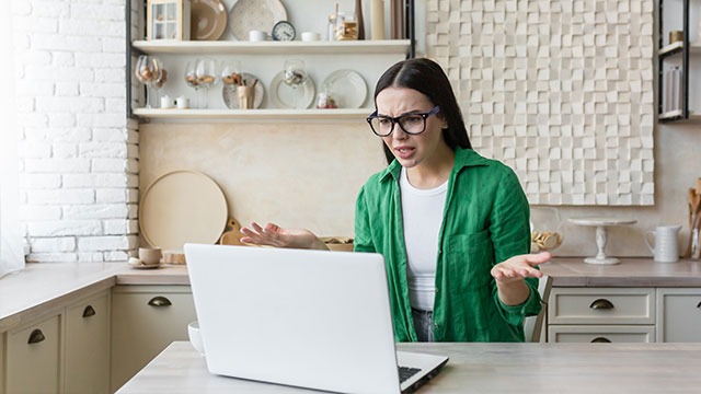 Een witte laptop staat op een tafel in een keuken met witte kasten en schappen met keukengerei.