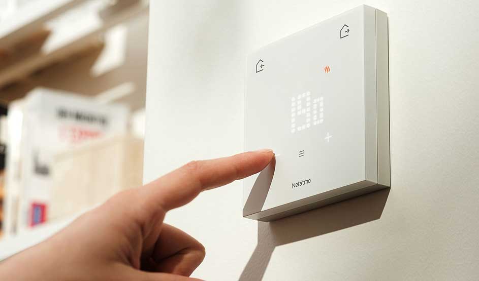Een vinger bedient een moderne witte thermostaat aan de muur, met digitaal display en aanraakknoppen.