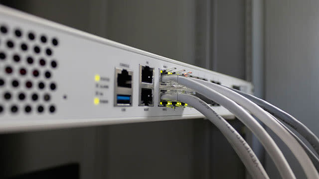 Een witte netwerkswitch met aangesloten grijze kabels tegen een grijze achtergrond, met gele lampjes.