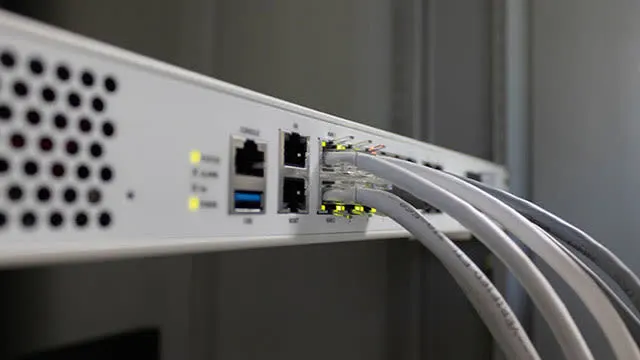 Een witte netwerkswitch met aangesloten grijze kabels tegen een grijze achtergrond, met gele lampjes.