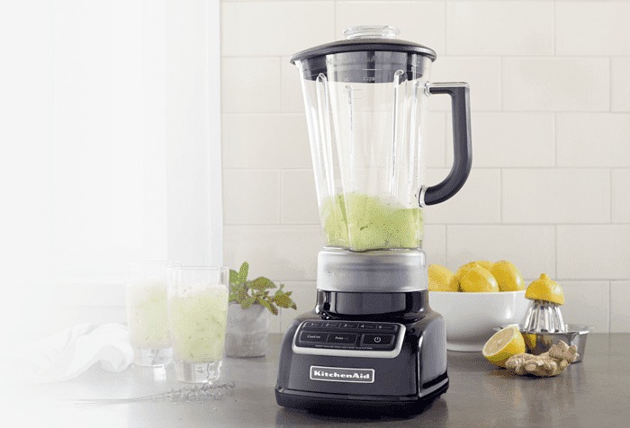 Un mélangeur noir KitchenAid remplit à moitié de liquide vert, avec des citrons, du gingembre et un presse-agrumes à proximité.