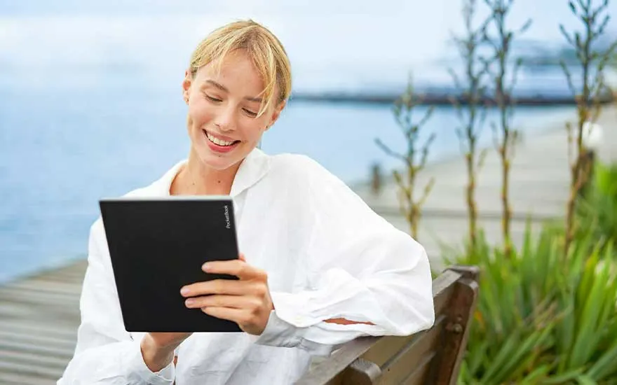 Een zwart tablet wordt vastgehouden tegen een achtergrond van water en groen, zittend op een houten bank.