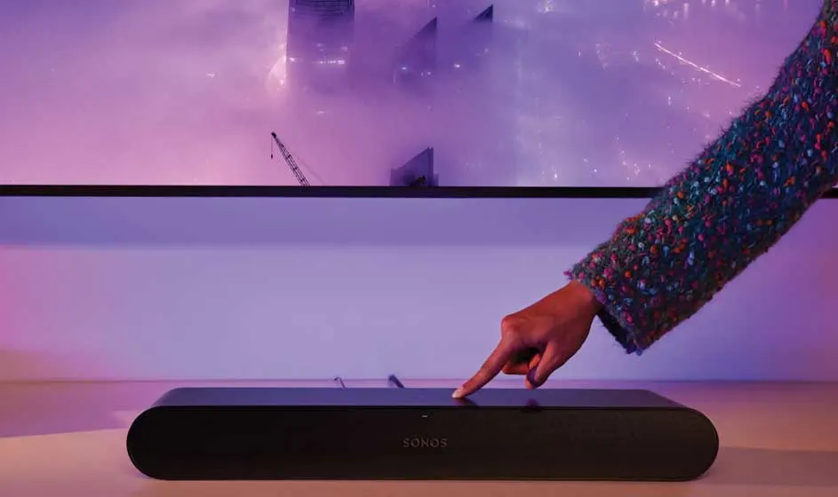 Une main touche une barre de son noire placée sous un grand écran. Des lumières violettes et roses donnent à la pièce moderne une ambiance futuriste.