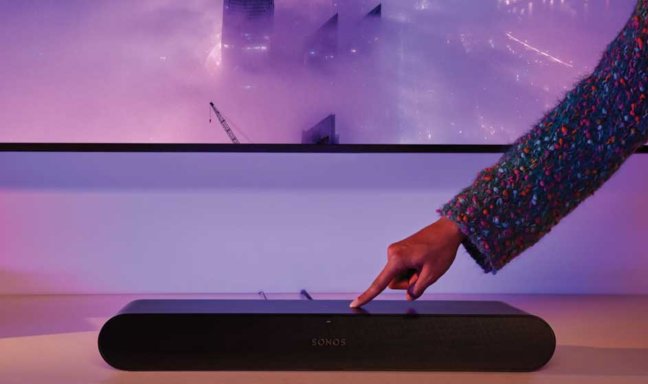 Een hand raakt een zwarte soundbar aan die onder een groot scherm staat. Paars en roze licht vullen de moderne kamer met een futuristische sfeer.