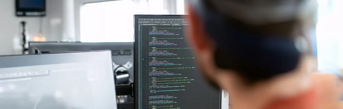 Un écran d'ordinateur affiche du code. Un autre écran, blanc, est visible à gauche. L'arrière-plan est clair et moderne.