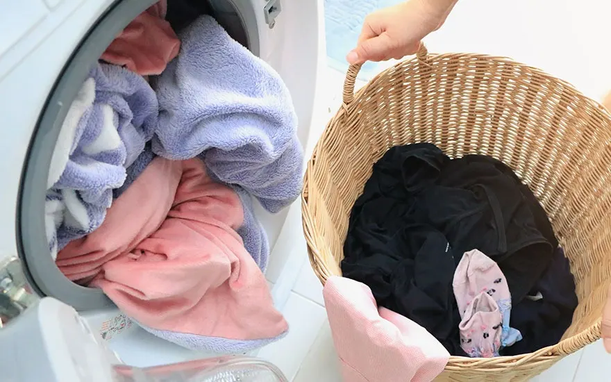 Un panier en osier rempli de linge noir, à côté d'un lave-linge rempli de linge rose et mauve.