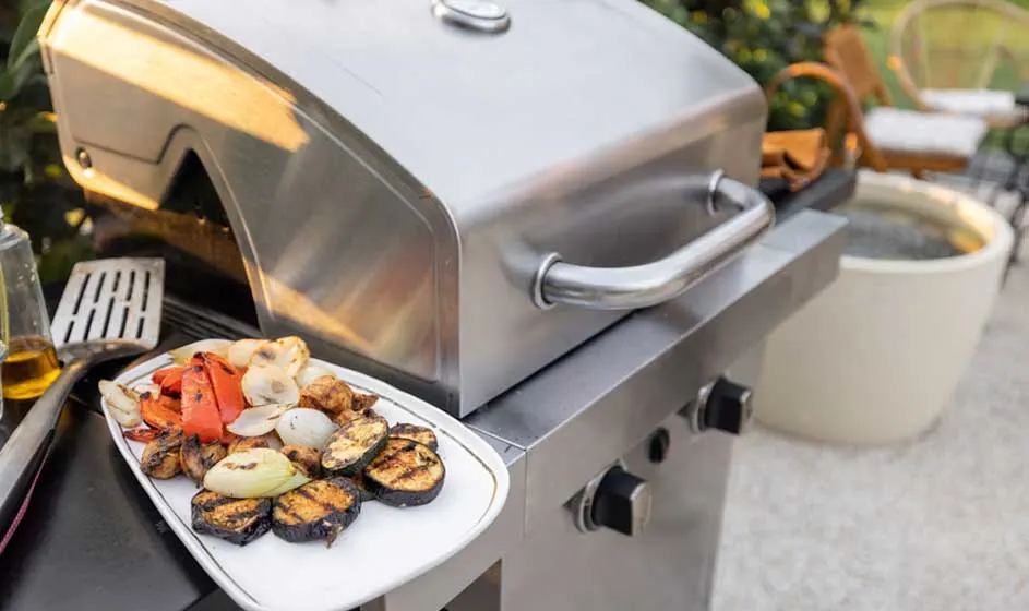 Een zilverkleurige barbecue met gegrilde groenten op een wit bord, met aubergine, paprika en ui.