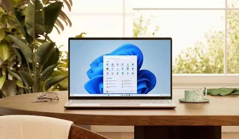 Laptop on a wooden table displays the Windows 11 interface, with a blue background. A cup, glasses, and a green plant are nearby.