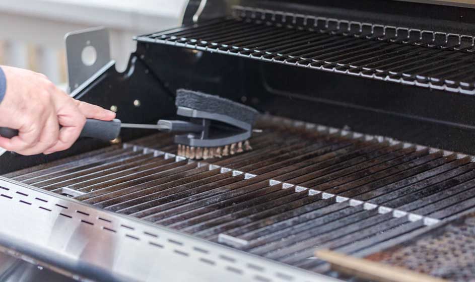 Een hand reinigt een zwarte grillrooster met een borstel. De grill is zilverkleurig en heeft horizontale staven.