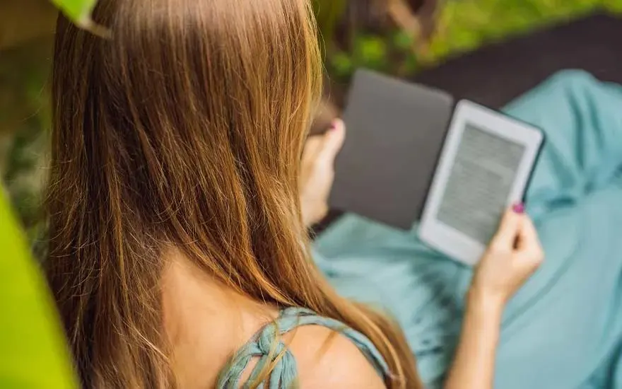 Une liseuse électronique blanche est tenue sur un tissu bleu clair, avec des feuilles vertes floues en arrière-plan.