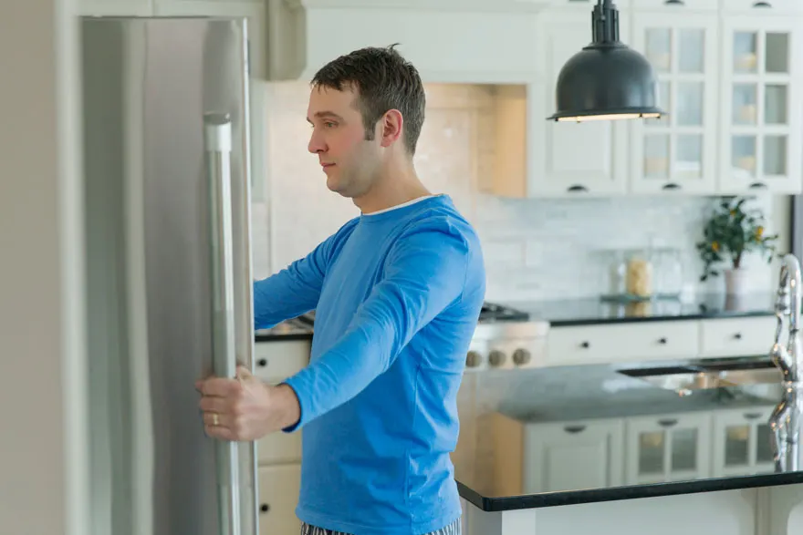 Un homme ouvre un frigo en acier dans une cuisine moderne blanche et noire. Une lampe pend au plafond.