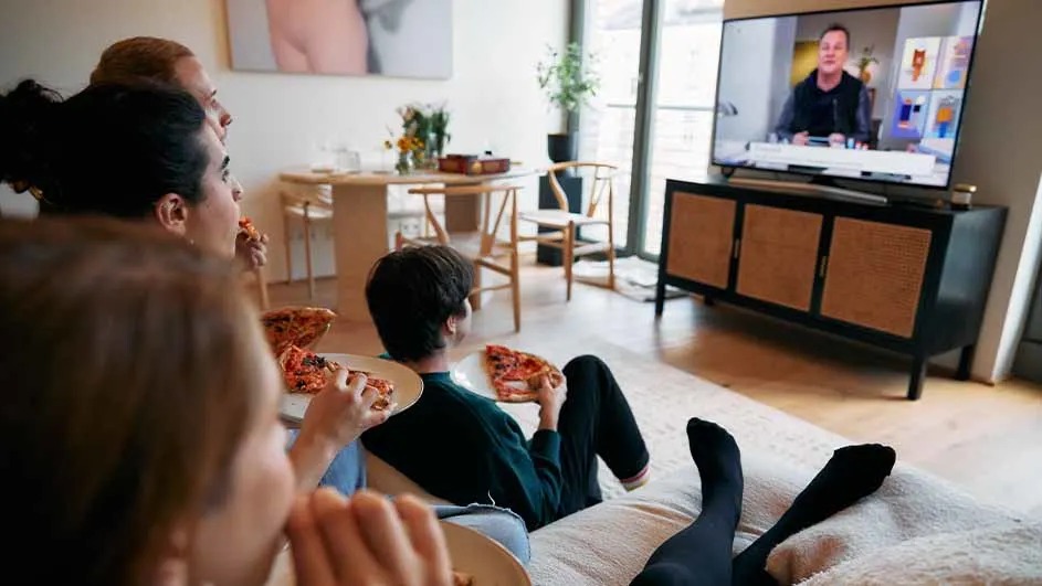 Un groupe mange de la pizza devant une télé. Des meubles en bois et une plante sont visibles en arrière-plan.