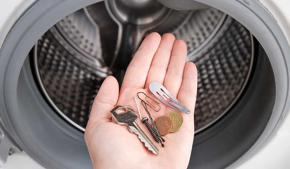 A hand holds found items like a key, hairpin, coins and a paperclip in front of an open washing machine. No brand is visible.