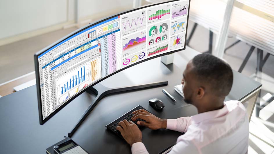 Zwarte bureau met breedbeeldscherm toont grafieken en spreadsheets. Toetsenbord, muis, notitieboek en rekenmachine zijn zichtbaar.