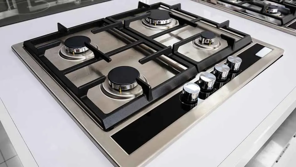 Table de cuisson à gaz en acier inoxydable avec quatre brûleurs noirs et quatre boutons de commande chromés sur un comptoir blanc.