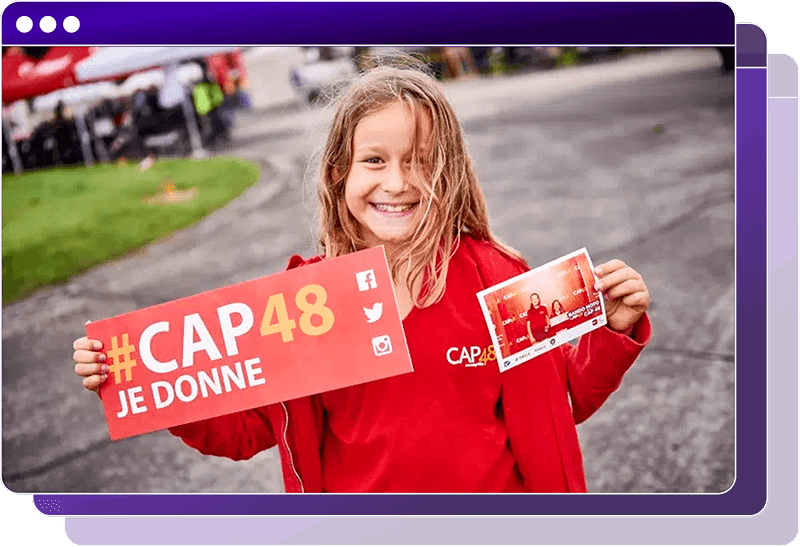 Person in a red CAP48 shirt holds a sign reading “#CAP48 JE DONNE” with social media icons and shows a small photo, outdoors at an event.