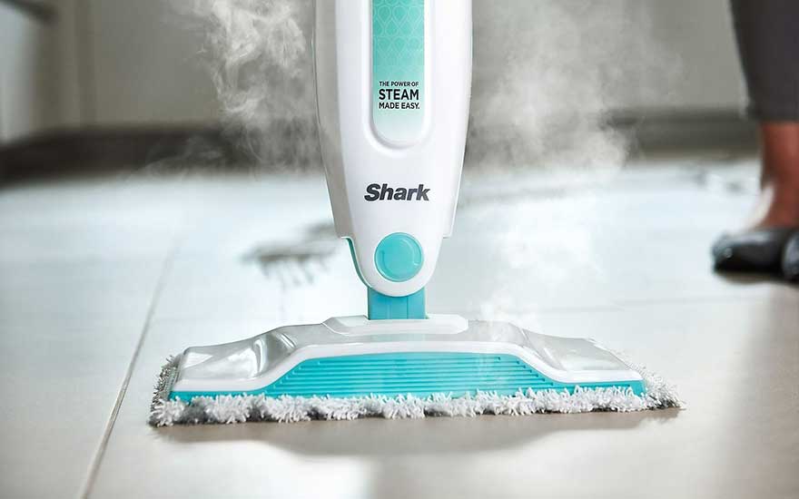Un nettoyeur vapeur blanc et turquoise "Shark" nettoie un sol carrelé, de la vapeur s'échappe de l'appareil.