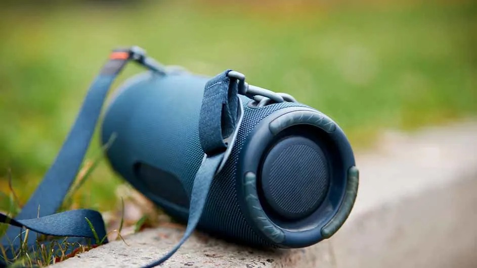 Haut-parleur bleu cylindrique avec sangle, posé sur une surface en béton devant un fond vert flou.