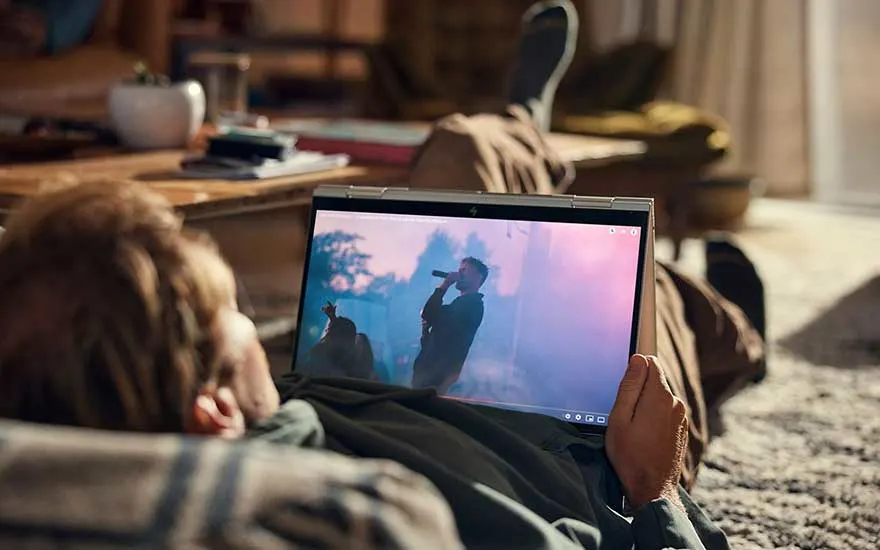 Een laptop toont een concert. De achtergrond is wazig. Op de achtergrond een houten tafel met een witte potplant.