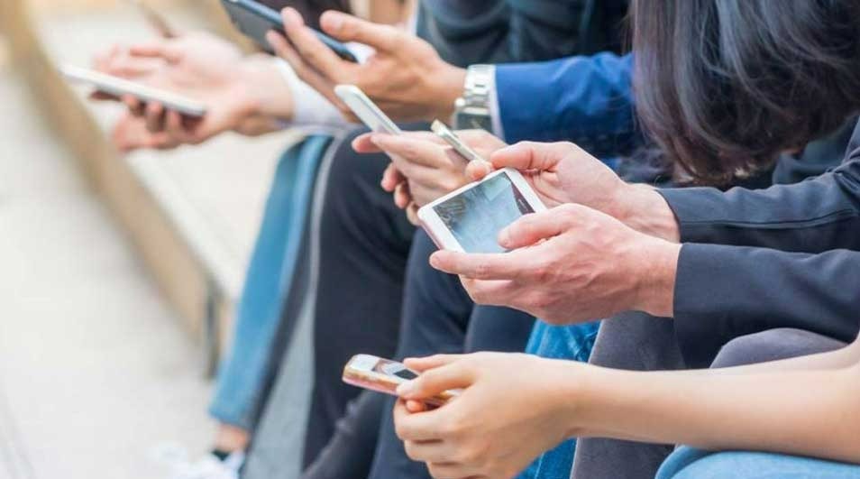 Several hands hold various smartphones and tablets, showing screens. Clothing includes blue jackets and jeans.