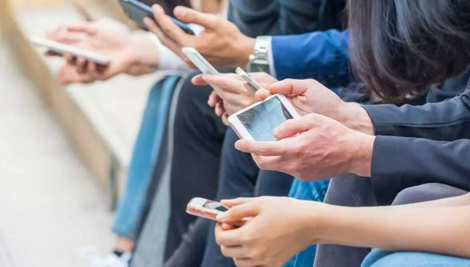 Several hands hold various smartphones and tablets, showing screens. Clothing includes blue jackets and jeans.