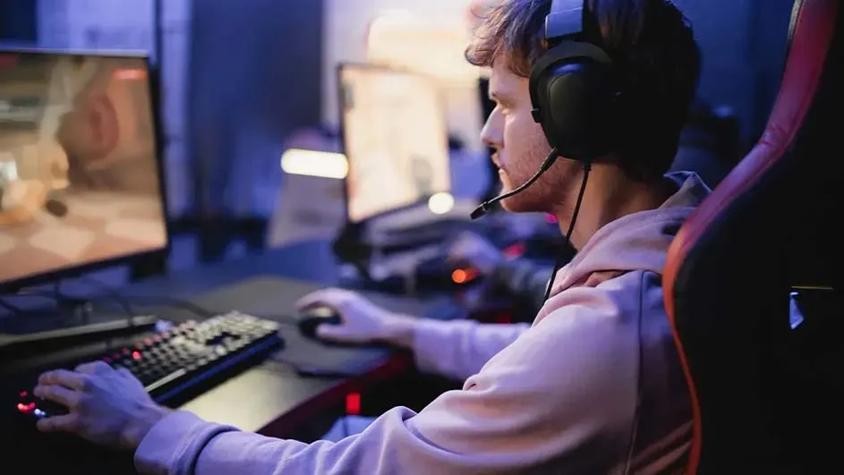 Een persoon met een headset speelt op een computer met een toetsenbord en muis in een blauwe kamer. Er staan meerdere schermen.