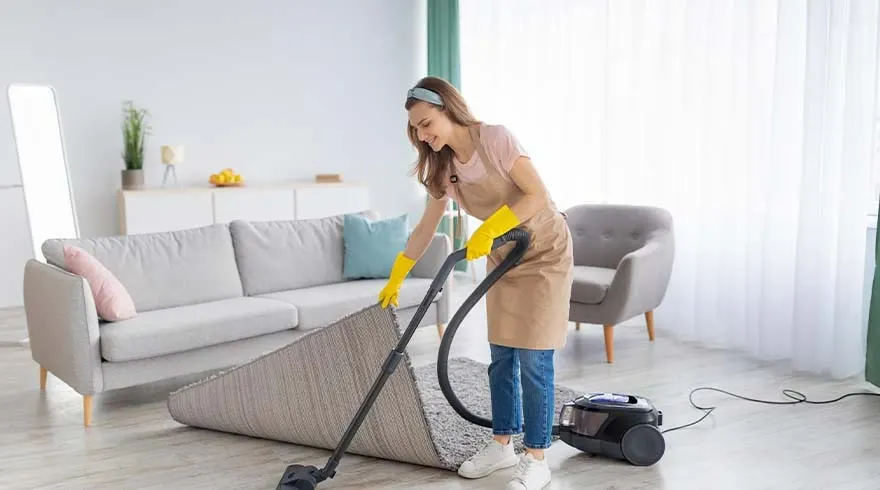 Woonkamer met grijze zetel, stoel en een beige tapijt dat gestofzuigd wordt. Een zwarte stofzuiger staat op de houten vloer.