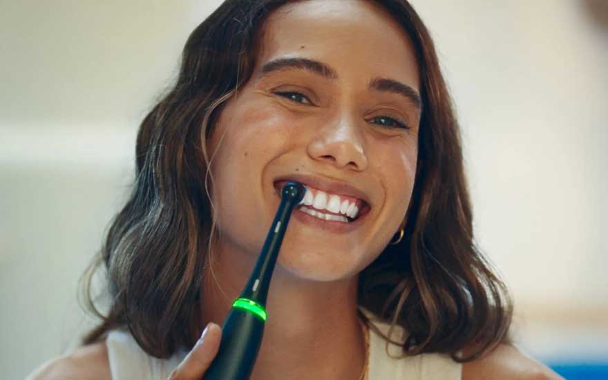 Une jeune femme se brosse les dents avec une brosse à dents électrique verte, tout en souriant largement devant la caméra.
