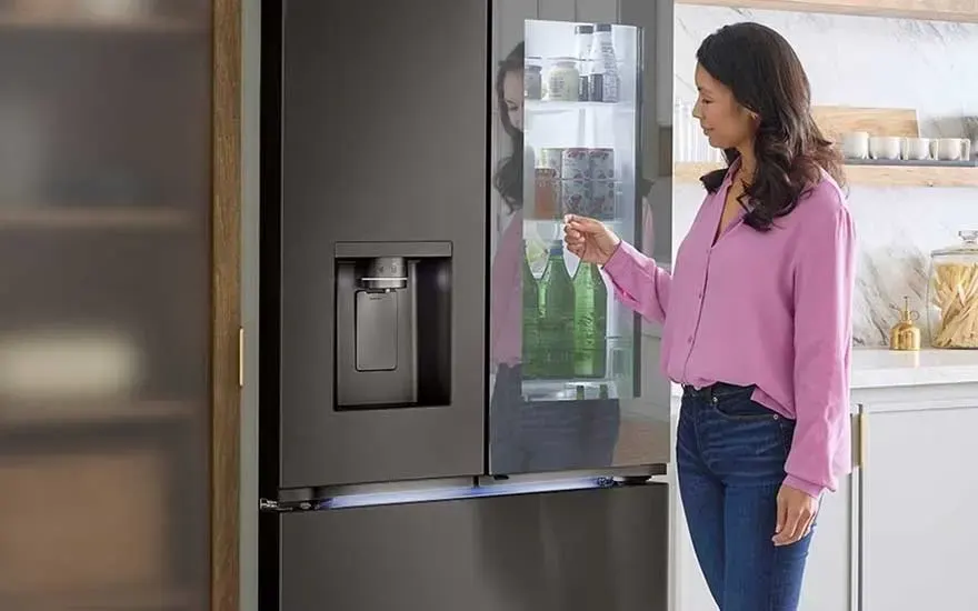 Een vrouw in een roze blouse staat bij een donkere koelkast met waterdispenser en bekijkt het verlichte scherm in de deur.