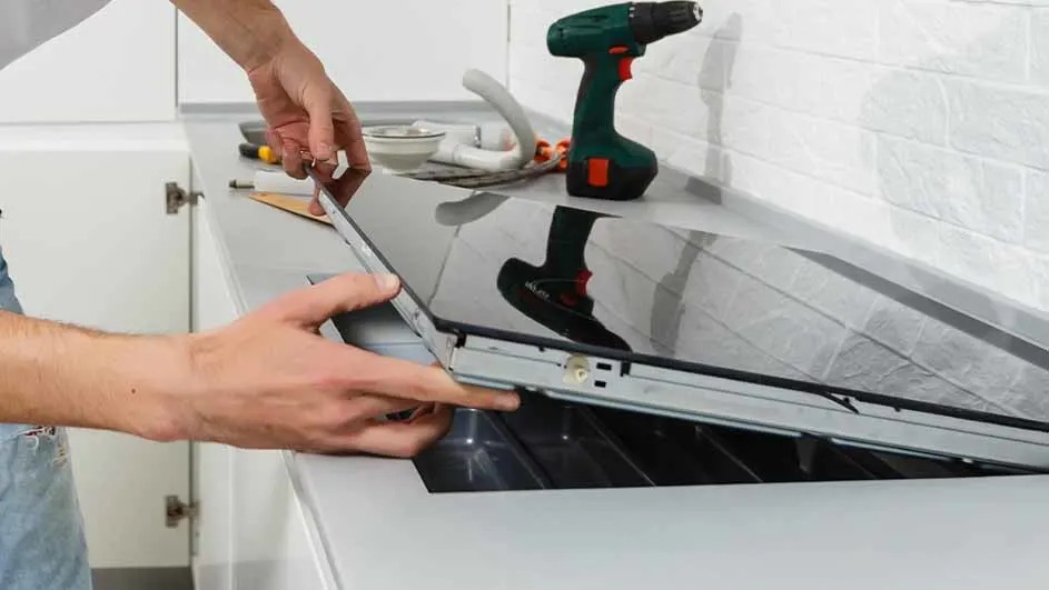 Un homme installe une plaque de cuisson noire sur un comptoir blanc, avec une perceuse à côté.
