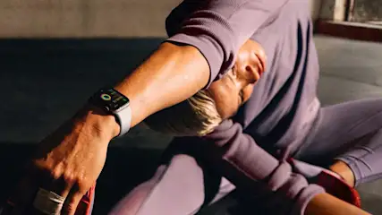 Een close-up van een horloge om een pols, met een lila trainingspak op de achtergrond en een grijze vloer.