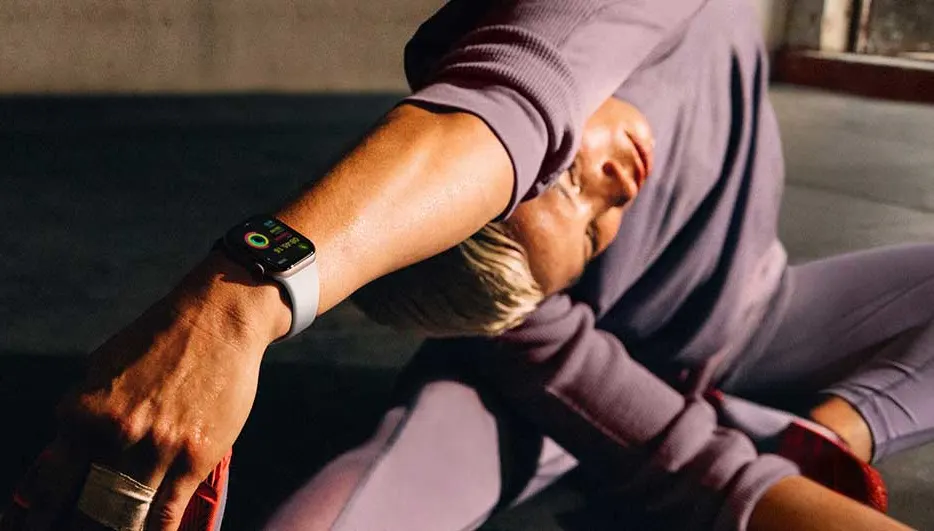 Een close-up van een horloge om een pols, met een lila trainingspak op de achtergrond en een grijze vloer.