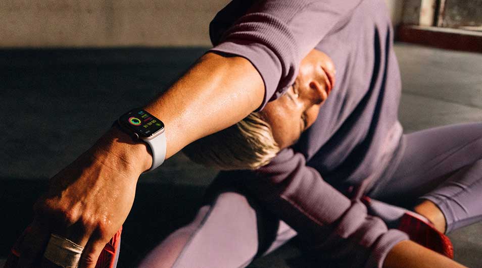 A fitness watch with a gray band is on a wrist in a purple long-sleeved shirt and leggings. The background is dark.
