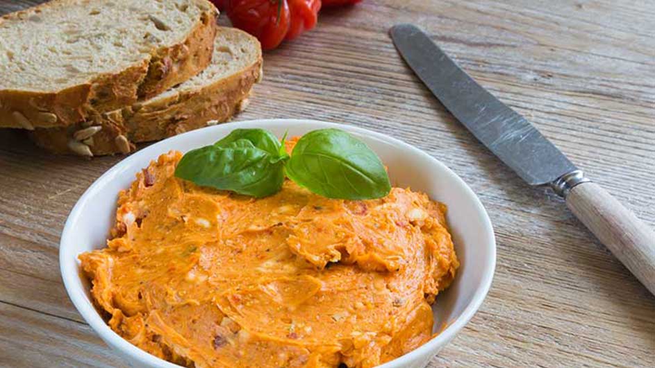 Un bol blanc de tartinade orange avec du basilic, du pain, des tomates et un couteau sur une table en bois.