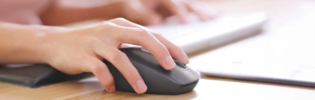 A hand operates a black wireless computer mouse on a wooden desk, with a keyboard blurred in the background.
