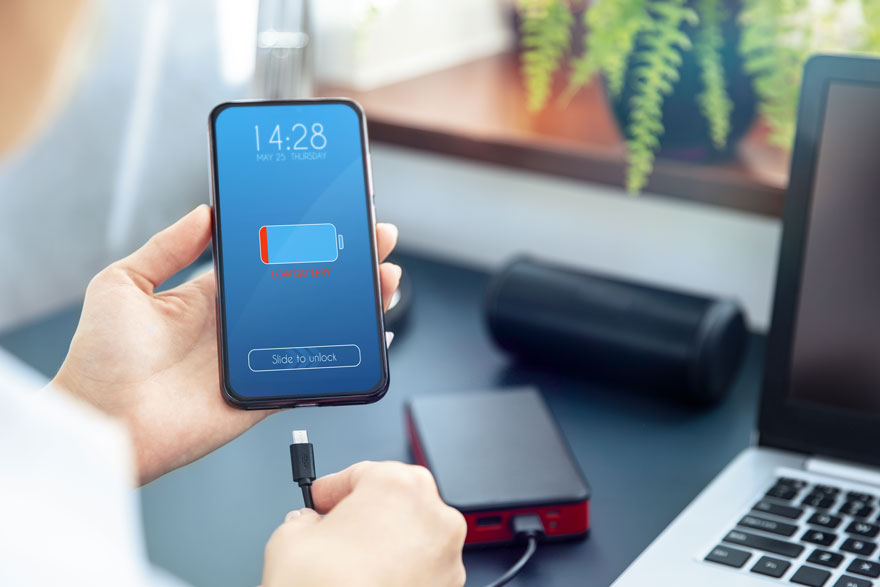 Een smartphone met blauw scherm toont een batterij bijna leeg. Iemand houdt een USB-kabel vast om op te laden. Op de achtergrond een laptop en speaker.
