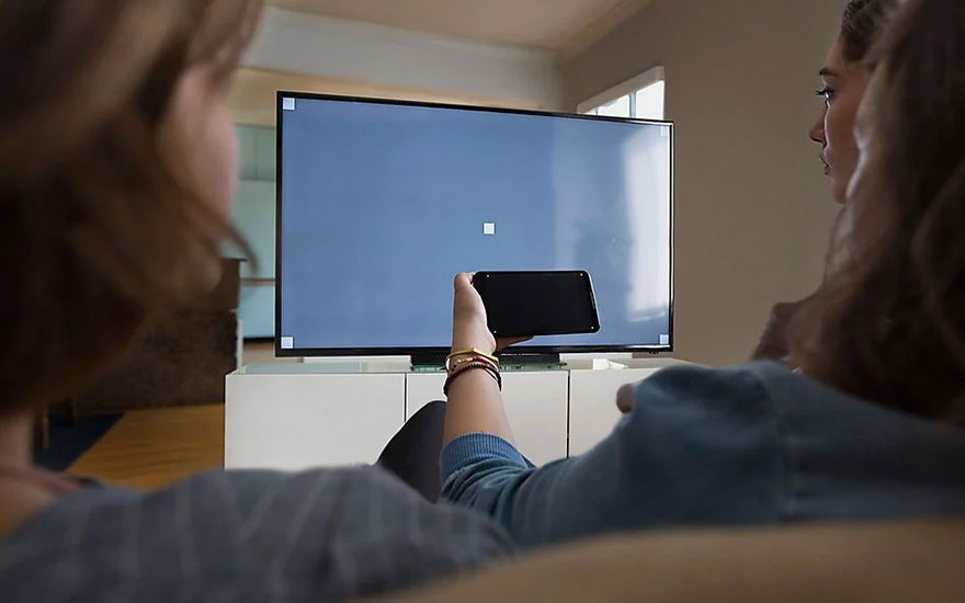 Deux personnes regardent une télévision à écran plat sur un meuble blanc. Une main tient un téléphone devant l'écran bleu.