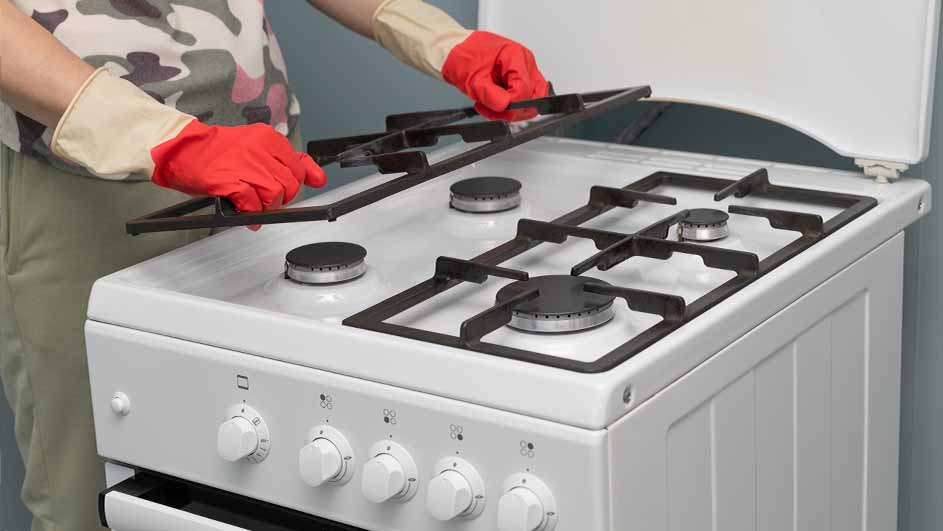A white stove with black grates is being cleaned. Red-gloved hands lift a grate above the stovetop. The background is a light gray wall.