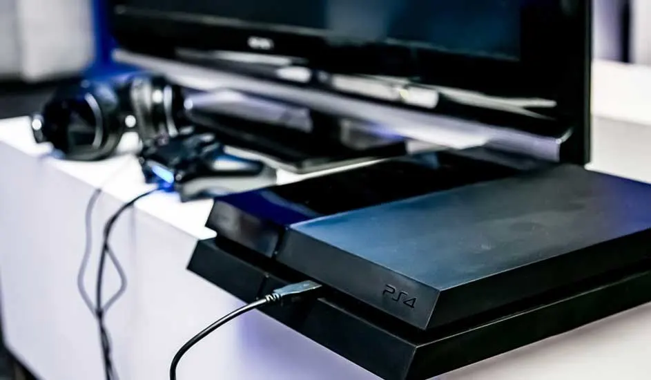 Een zwarte PlayStation 4 op een witte tafel, met een tv, koptelefoon en controller op de achtergrond.